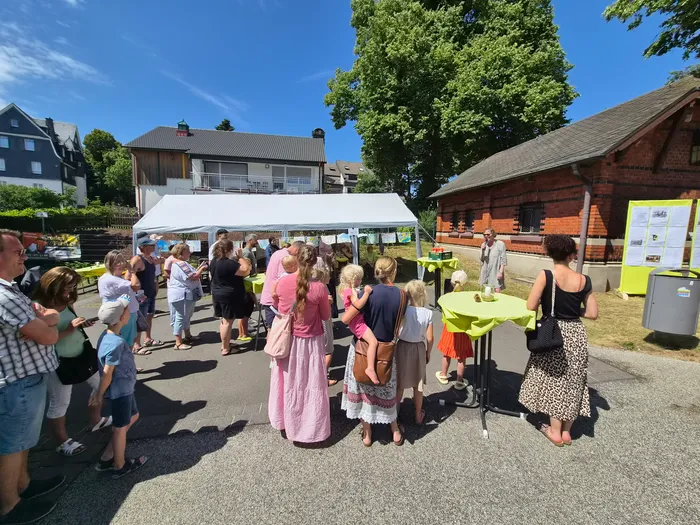 Menschen stehen und unterhalten sich auf einem Fest im Freien vor einem Zelt und Gebäuden. Im Hintergrund sind weitere Personen und grüne Bäume zu sehen.