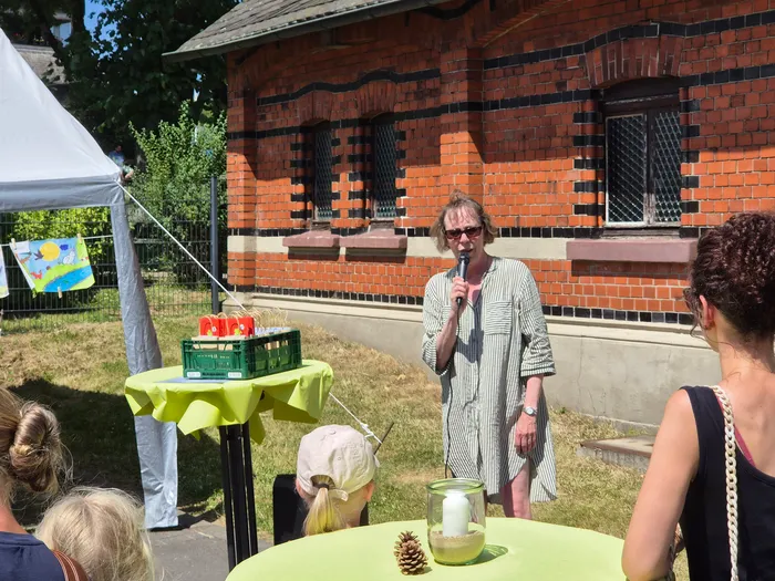 Eine Frau mit kurzem, blondem Haar hält ein Mikrofon in der Hand und spricht vor einer Gruppe von Menschen im Freien. Im Hintergrund ist ein rotes Backsteingebäude zu sehen.