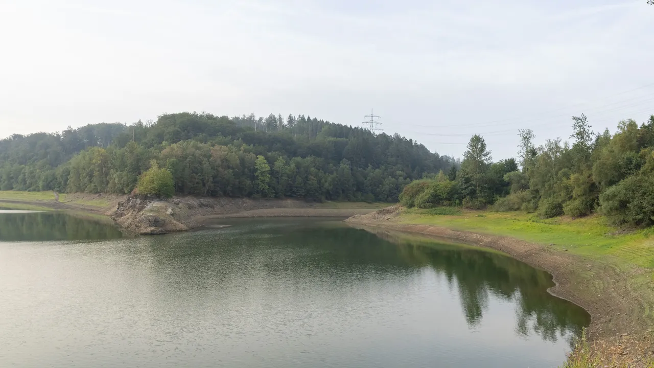 Die Wupper-Talsperre mit niedrigem Wasserstand und einem breiten, trockenen Uferstreifen vor bewaldeten Hügeln.