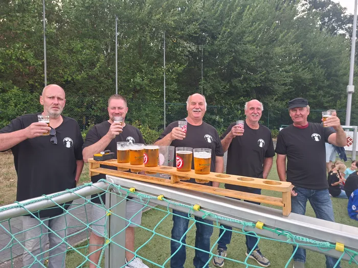 Fünf Männer in schwarzen T-Shirts halten beim Fußballturnier Gläser mit Bier hoch und stehen hinter einem hölzernen Biertisch.