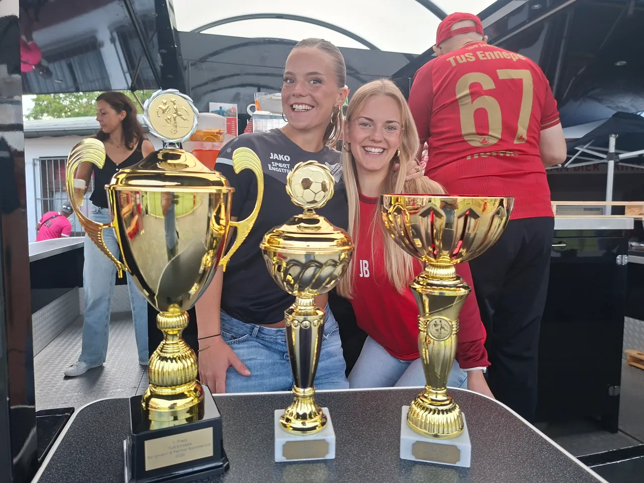 Zwei lächelnde junge Frauen posieren hinter drei goldenen Pokalen beim Hobby-Kicker-Turnier des TuS Ennepe.