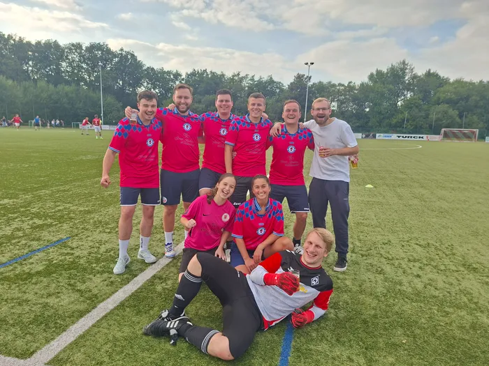 Eine Gruppe von Fußballspielern in rot-blauen Trikots posiert lachend auf einem Kunstrasenplatz.