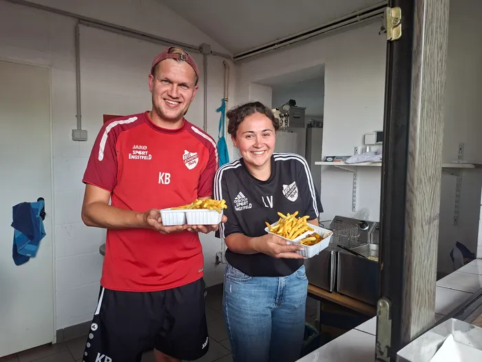 Ein Mann und eine Frau in Sportkleidung des TuS Ennepe halten lächelnd Schalen mit Pommes frites in die Kamera.