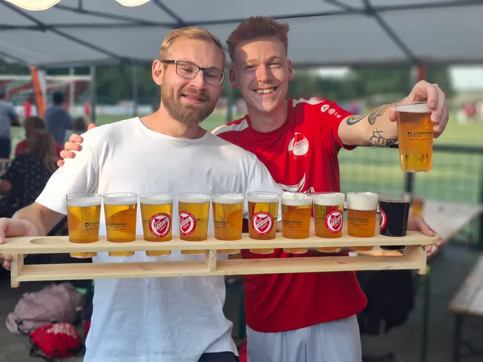 Zwei Männer halten ein Holztablett mit verschiedenen Biergläsern, einer von ihnen hebt ein Glas hoch.