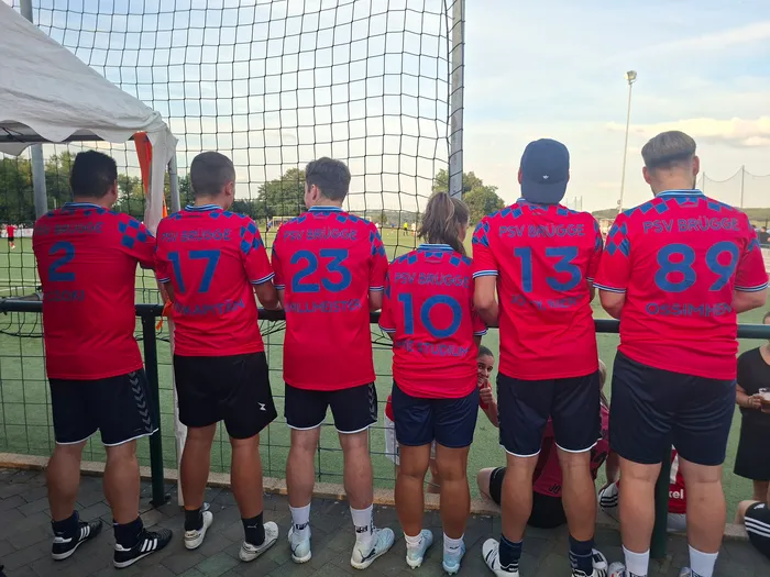 Sechs Fußballspieler in roten Trikots mit der Aufschrift „PSV BRÜGGE“ stehen mit dem Rücken zur Kamera vor einem Fußballtor.