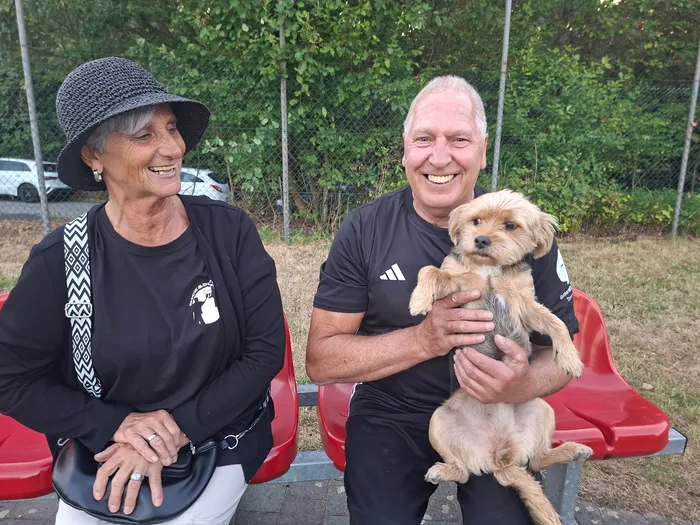Ein lächelnder Mann hält einen kleinen, hellbraunen Hund, während eine Frau mit Hut daneben sitzt.