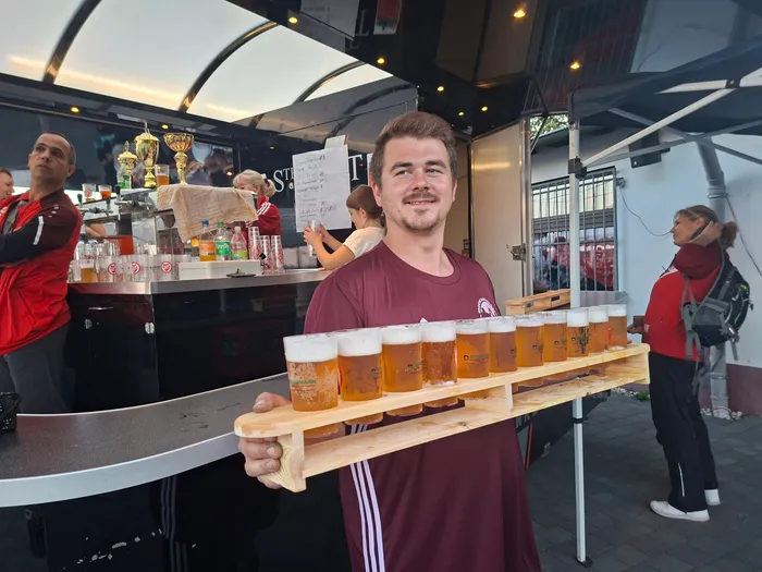 Ein Mann in einem weinroten T-Shirt hält ein Holztablett mit mehreren Gläsern Bier vor einem Verkaufsstand bei einem Fußballturnier.