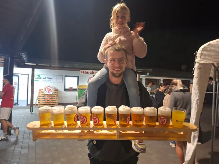 Ein lächelnder Mann trägt ein Mädchen auf den Schultern und hält ein großes Holztablett mit zehn gefüllten Biergläsern.