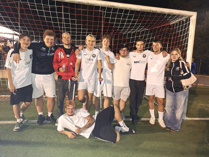 Eine Gruppe junger Fußballspieler und eine junge Frau posieren vor einem Fußballtor auf einem Rasenplatz bei Nacht.