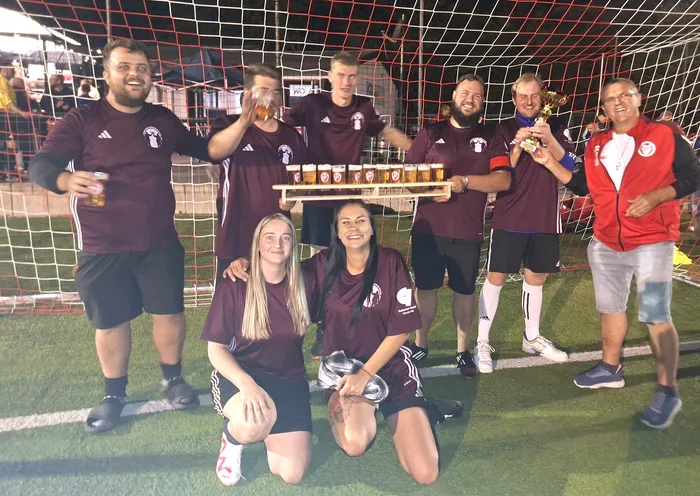 Eine Gruppe von Spielern in weinroten Trikots feiert vor einem Fußballtor mit einem Pokal und einem Tablett voller Gläser.