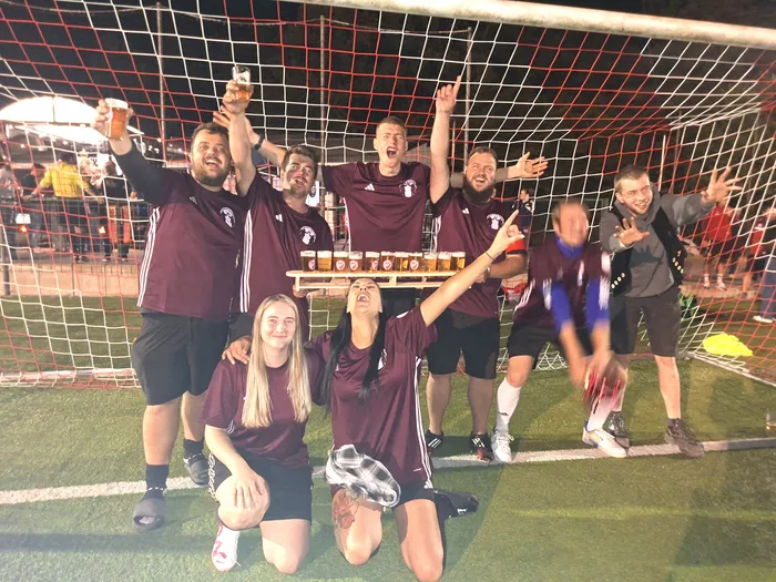 Eine Gruppe jubelnder Menschen in weinroten Sporttrikots feiert vor einem Fußballtor mit Biergläsern bei Nacht.