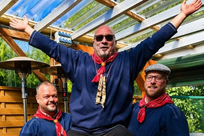 Colin Storm mit erhobenen Armen und Sonnenbrille, flankiert von zwei Männern in blauen Hemden und roten Halstüchern.