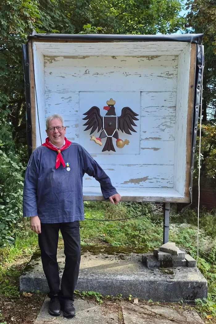 Norbert Falkenroth steht neben einer großen weißen Holzkonstruktion mit einem hölzernen schwarzen Adler und einem Wappenschild.