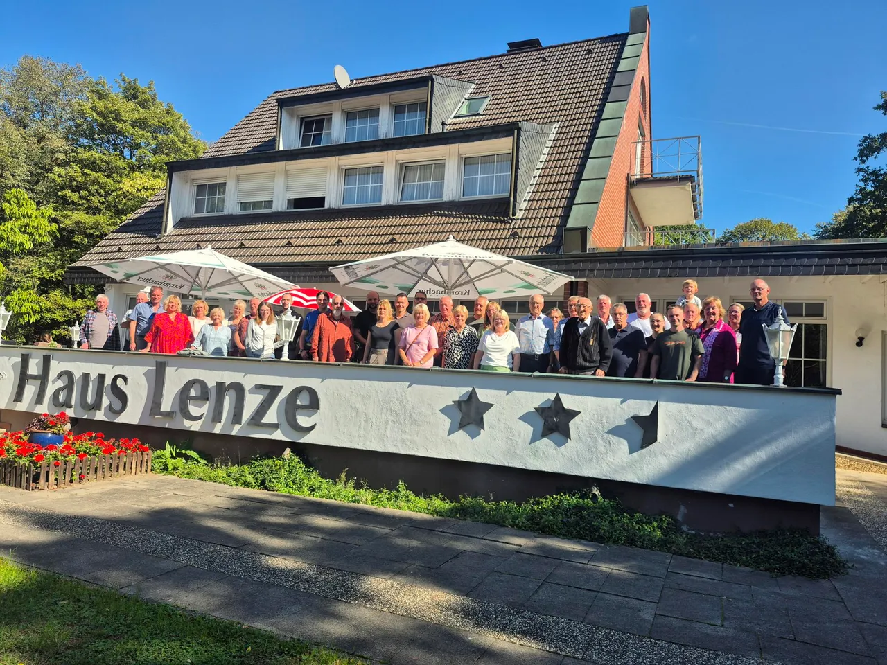 Eine große Gruppe von Menschen posiert vor dem Gebäude „Haus Lenze“ unter weißen Sonnenschirmen.