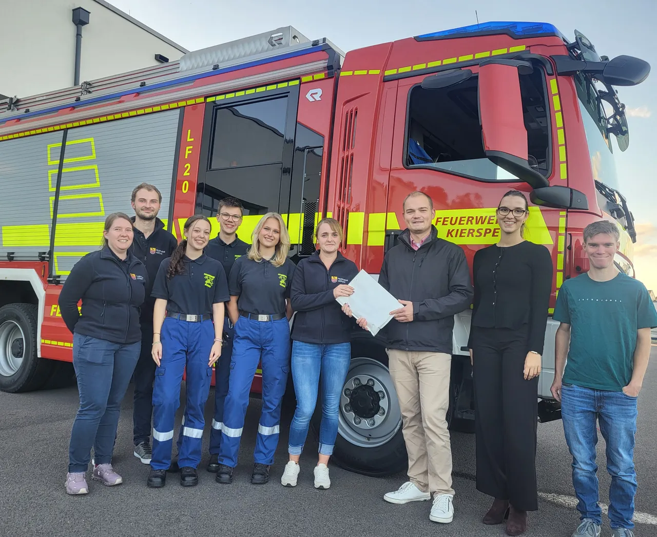 Lennart Wüst überreicht Anica Dörseln einen weißen Umschlag vor einem roten Feuerwehrfahrzeug der Feuerwehr Kierspe.