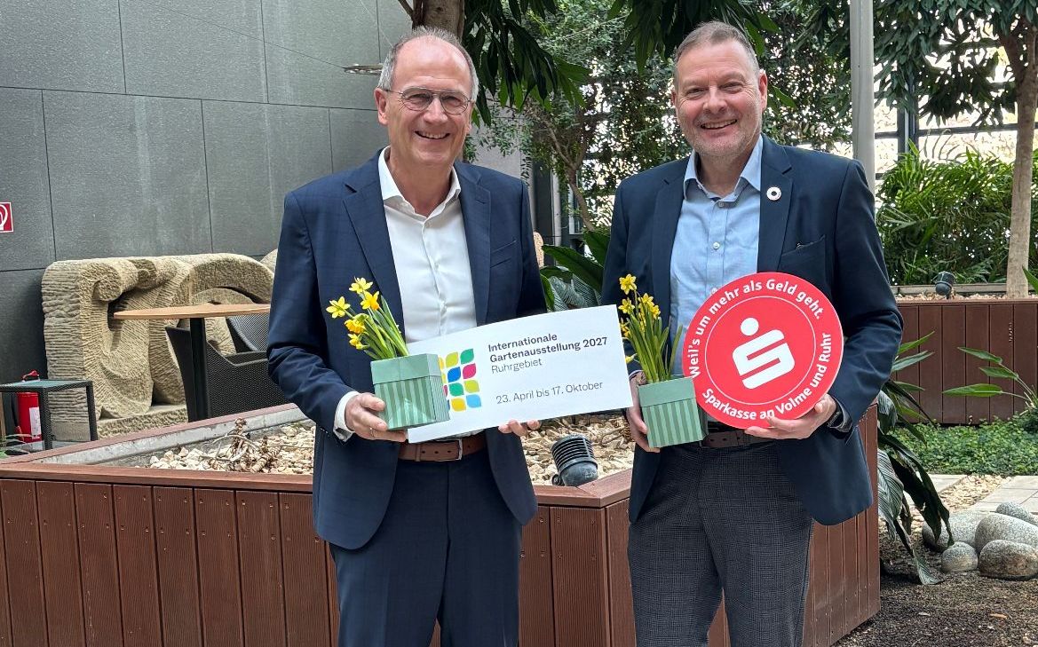 Zwei Männer in Anzügen stehen vor einem erhöhten Blumenbeet und halten ein Werbeschild für die Internationale Gartenausstellung 2027. Im Hintergrund ist eine grüne Pflanzenwand zu sehen.