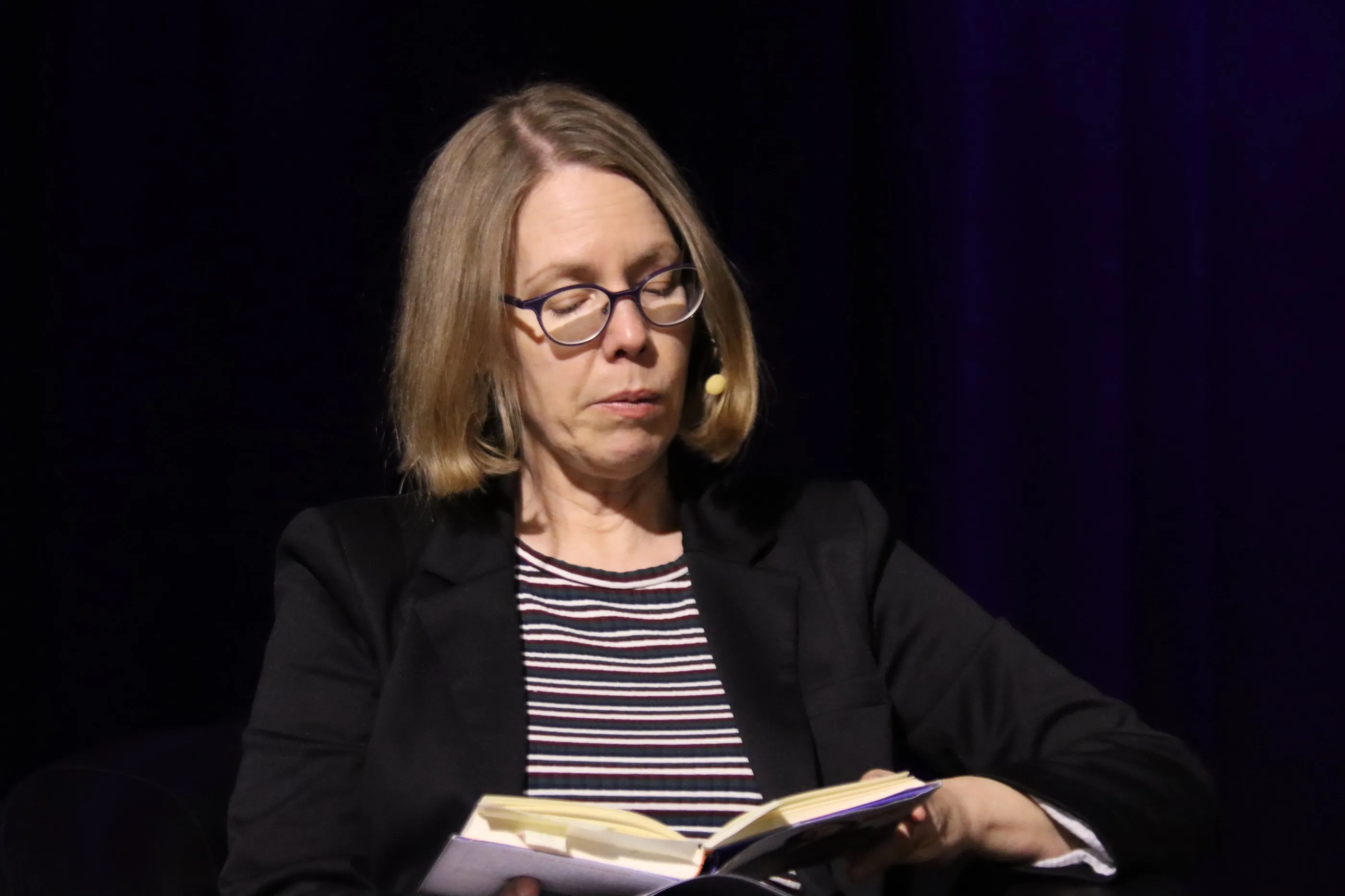 Frau mit Brille liest von Notizen ab, während sie auf einer Bühne spricht. Sie trägt einen Blazer über einem gestreiften Oberteil.