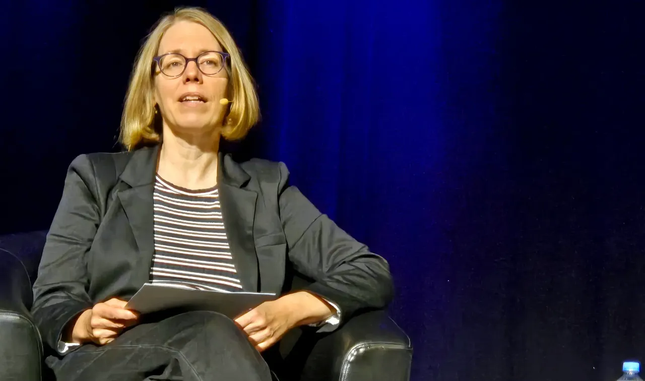 Anne Brorhilker sitzt auf einem Ledersessel, hält ein Blatt Papier und spricht in ein Headset. Im Hintergrund ein dunkler Vorhang und eine Flasche Wasser.
