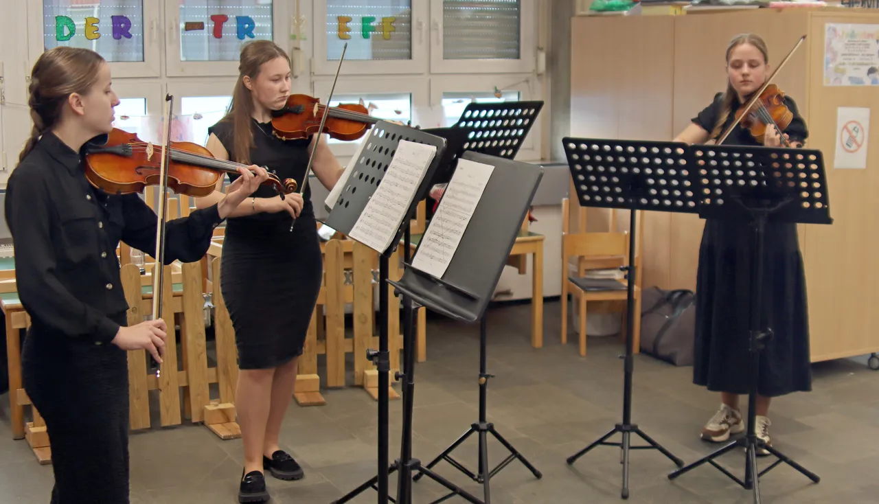 Drei junge Frauen spielen Violine vor Notenständern in einem hellen Raum, im Hintergrund ein Fenster und bunte Buchstaben.