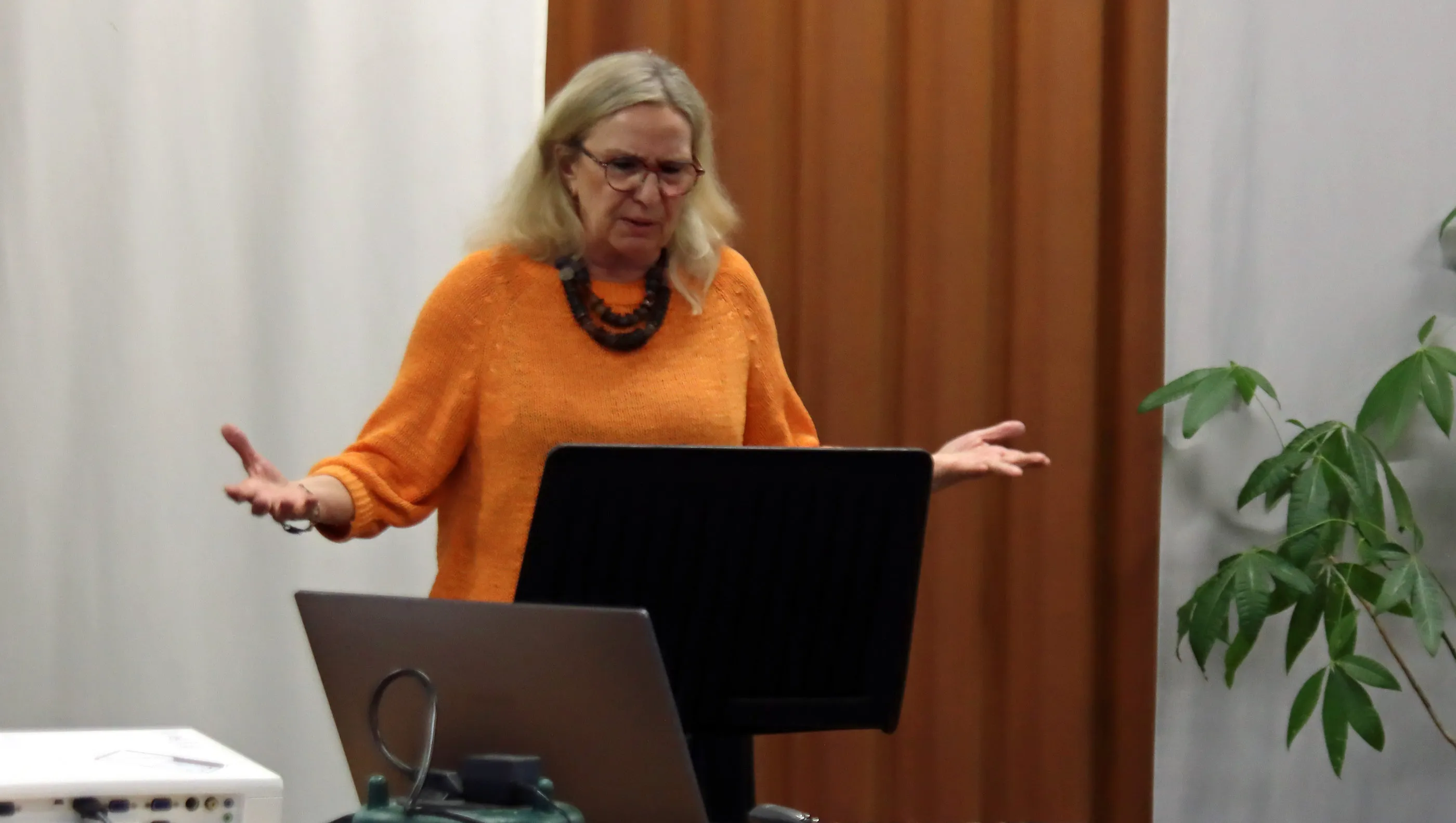 Eine Frau mit blonden Haaren und Brille steht vor einem Laptop und einem schwarzen Rednerpult, die Hände sind erhoben und sprechen. Im Hintergrund ein orangefarbener Vorhang und eine Pflanze.