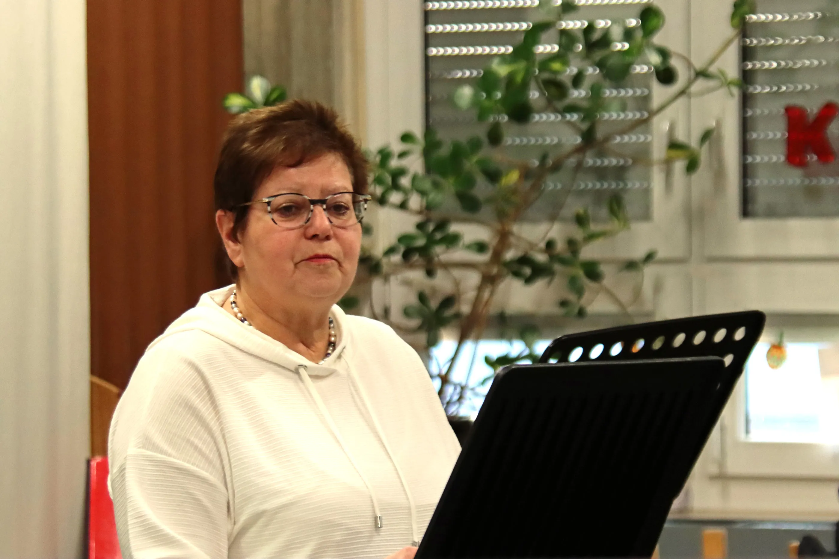 Eine Frau mit kurzem braunem Haar und Brille liest von einem Notenständer, im Hintergrund eine Pflanze und ein Fenster.