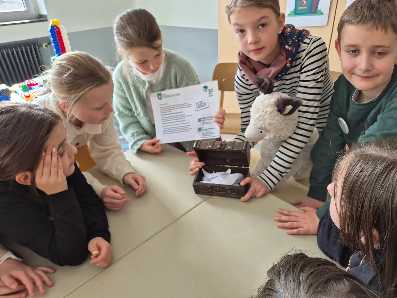 Eine Gruppe von Kindern sitzt um einen Tisch und betrachtet eine geöffnete Schatztruhe, aus der Papierstücke herausragen. Ein Kind hält ein Arbeitsblatt und ein Stofftier in der Hand.