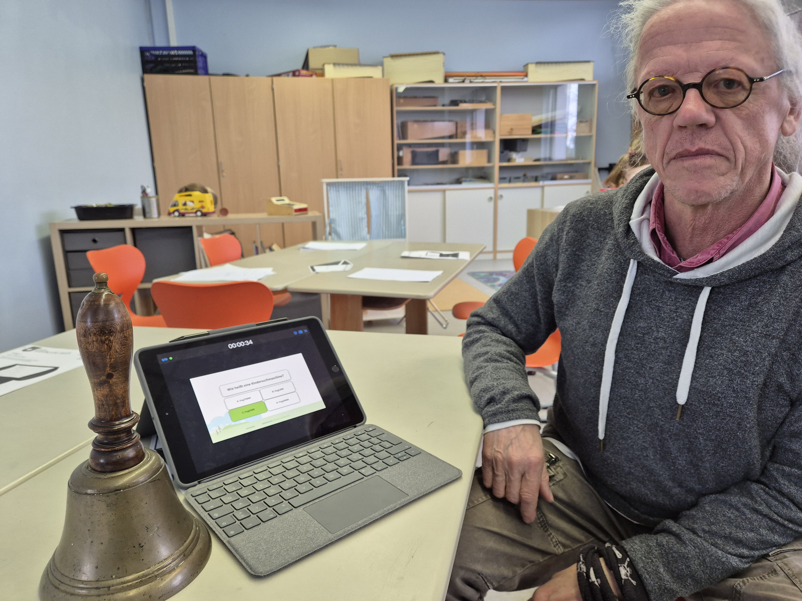 Ein älterer Mann mit Brille und grauen Haaren lächelt in die Kamera, während er neben einem Laptop und einer alten Schulglocke sitzt. Im Hintergrund sind Tische und Stühle in einem Klassenzimmer zu sehen.