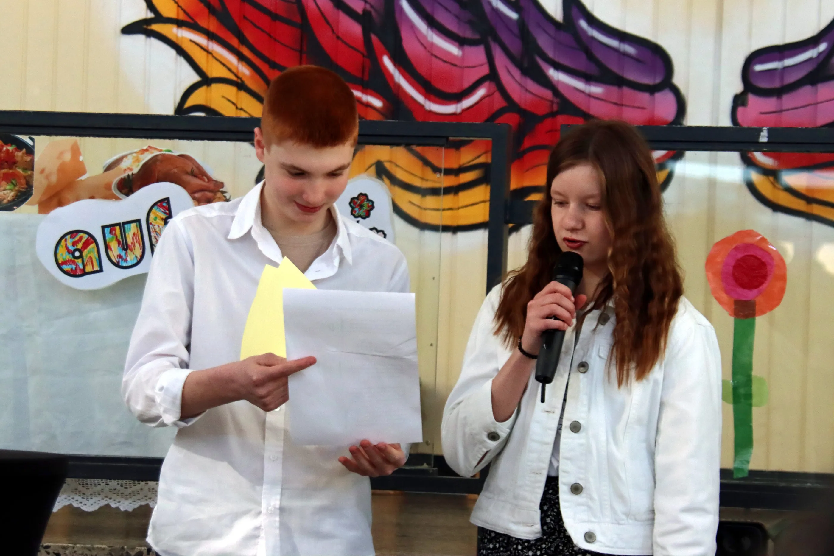 Zwei Jugendliche stehen auf einer Bühne. Der Junge hält ein Blatt Papier und zeigt darauf, während das Mädchen in einen Mikrofon spricht. Im Hintergrund eine bunte Wandgestaltung mit Blumen und Lebensmitteldarstellungen.