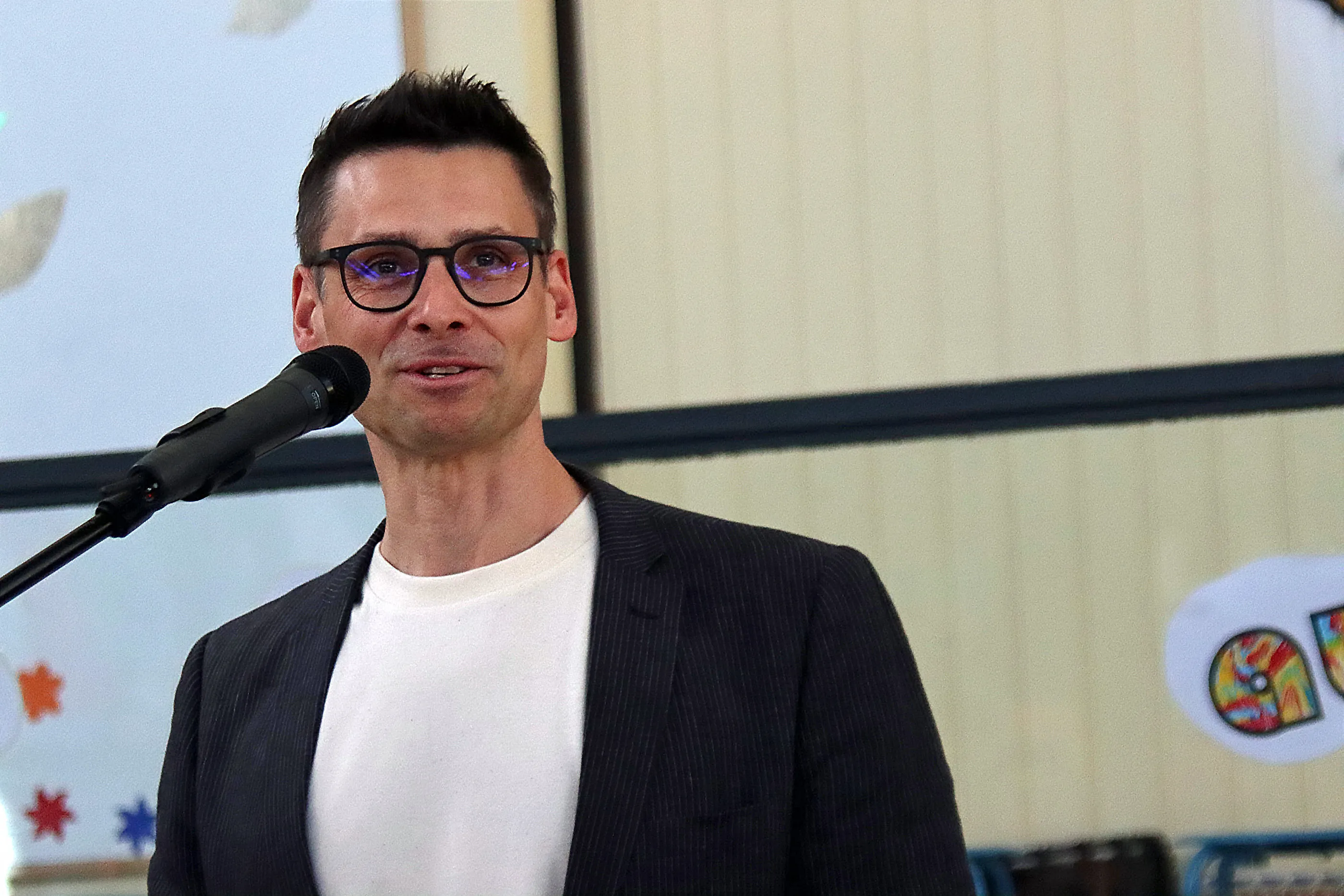 Mann mit Brille spricht in ein Mikrofon auf einem Frühlingsfest. Im Hintergrund sind bunte Dekorationen zu sehen.