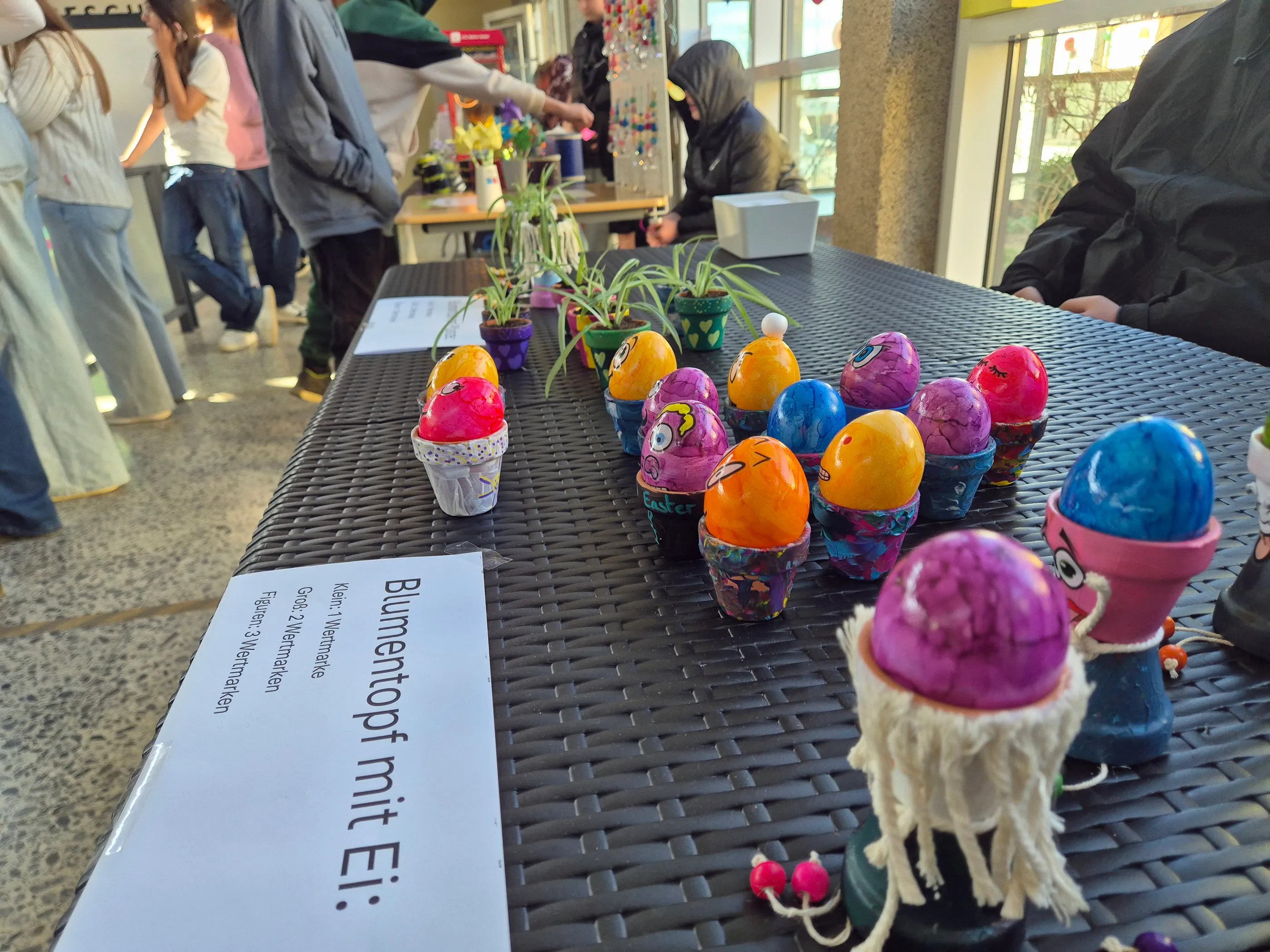 Bunte Ostereier stehen auf einem Tisch neben kleinen Blumengestecken. Im Vordergrund ein Blatt mit Preisinformationen für Blumentöpfe mit Eiern.