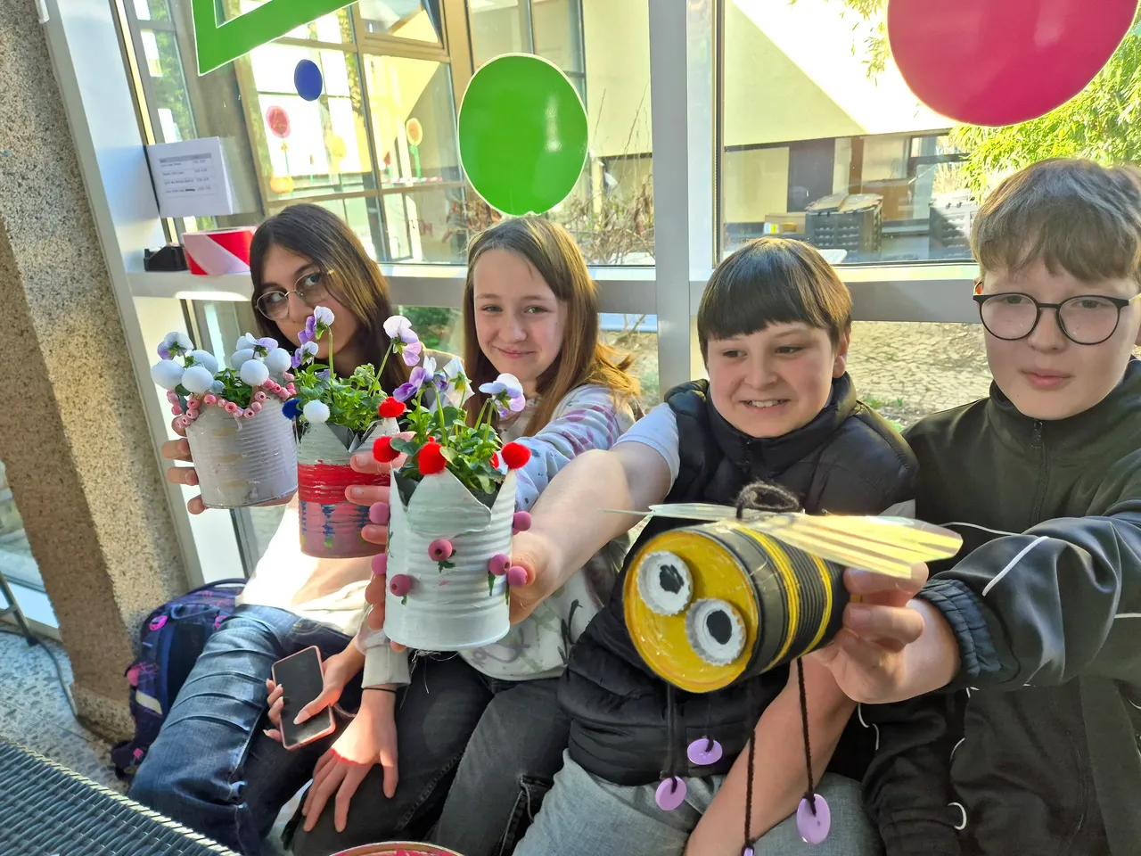 Vier Kinder präsentieren selbstgebastelte Frühlingsdekorationen aus Dosen und Blumen vor dem Eingang der Phönixschule.