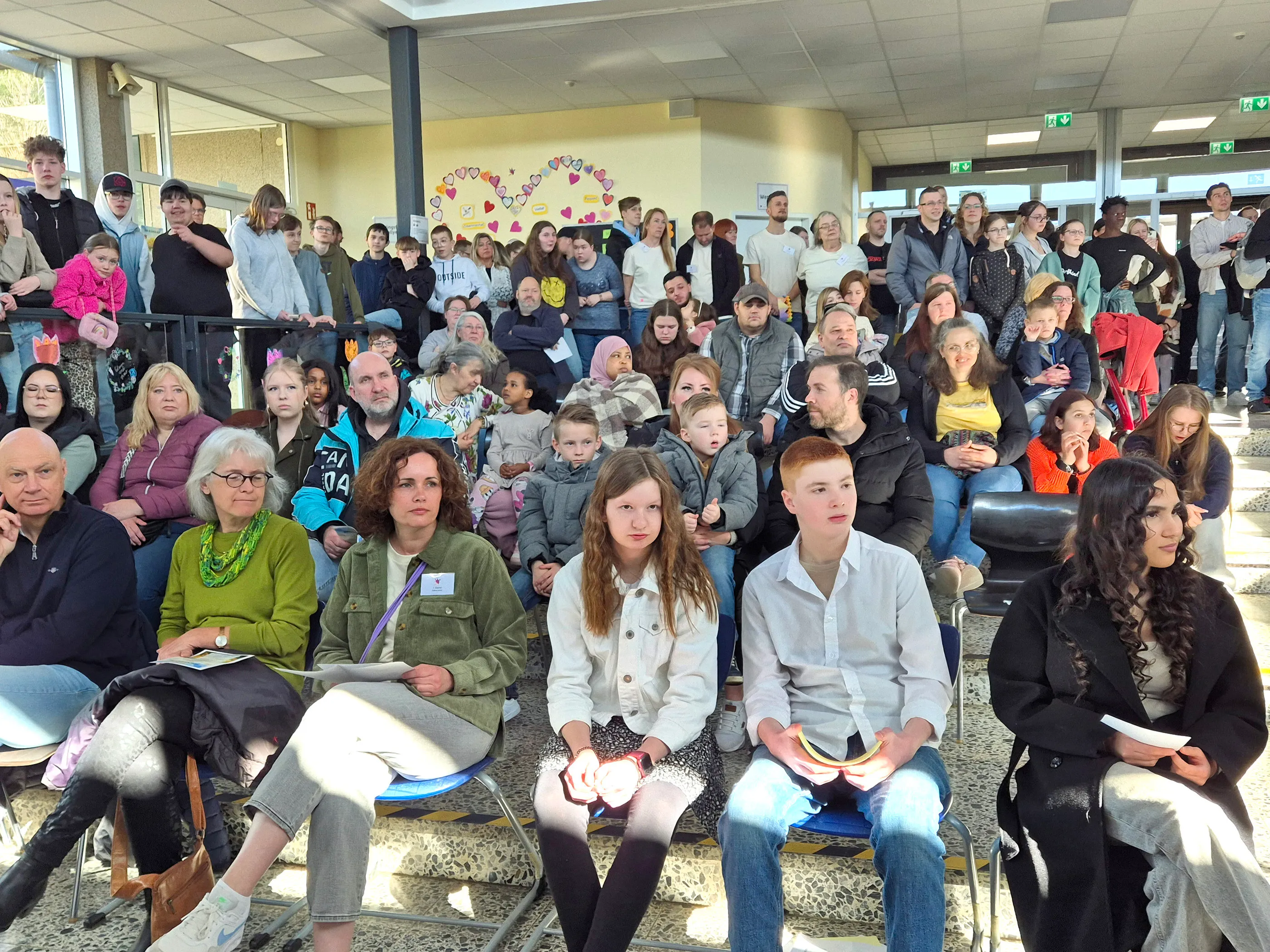 Große Menschengruppe sitzt und steht in einer hellen Halle mit Herzmotiven an der Wand, vermutlich bei einem Frühlingsfest der Phönixschule.