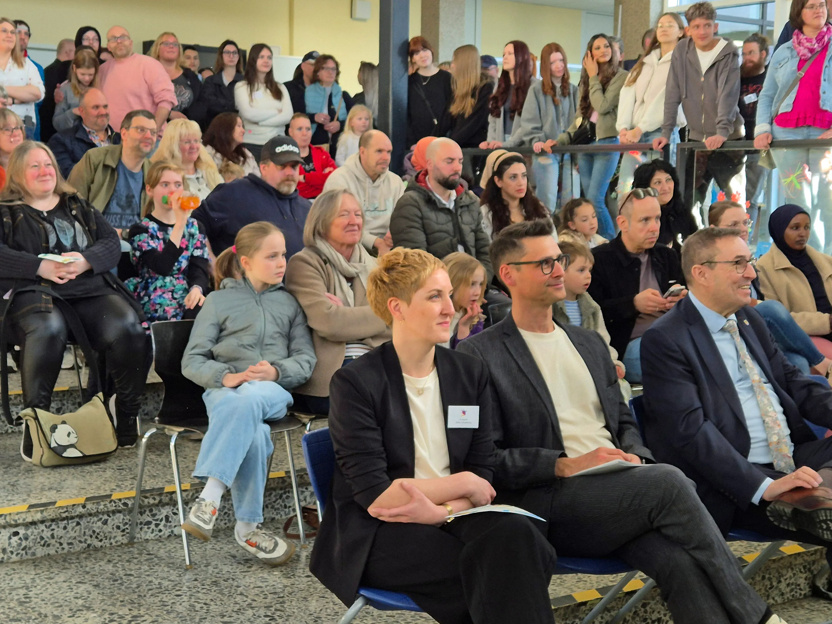 Zuschauer sitzen auf blauen Stühlen in einer hellen Halle, viele blicken in eine Richtung, vermutlich auf eine Bühne. Im Vordergrund eine Frau mit rotem Haar und ein Mann mit Brille im Anzug.