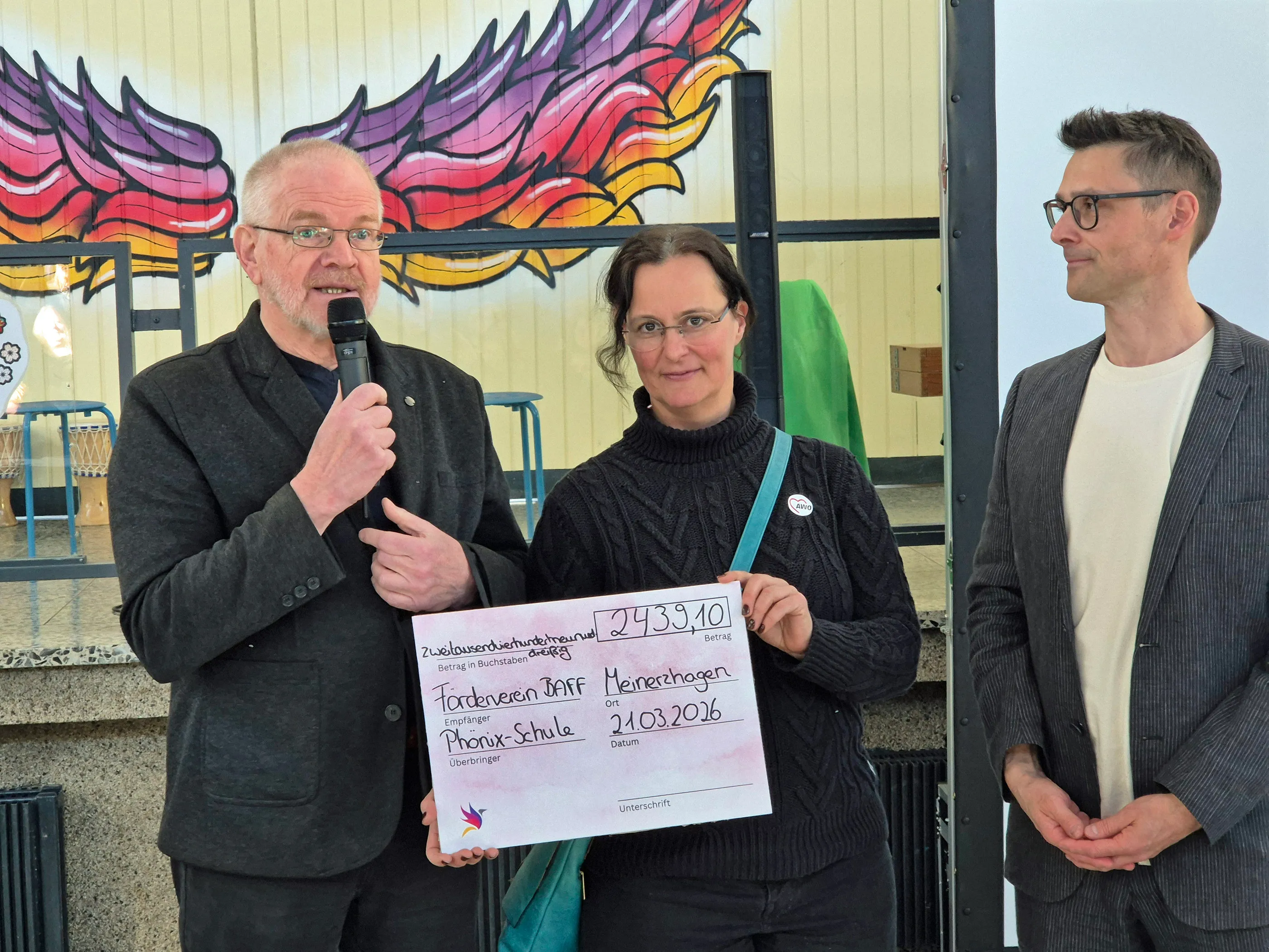 Zwei Männer überreichen einen Scheck über 2433,10 Euro an eine Frau vor einer bunten Wand. Der Scheck ist ausgestellt an die Phönix-Schule in Meinerzhagen.