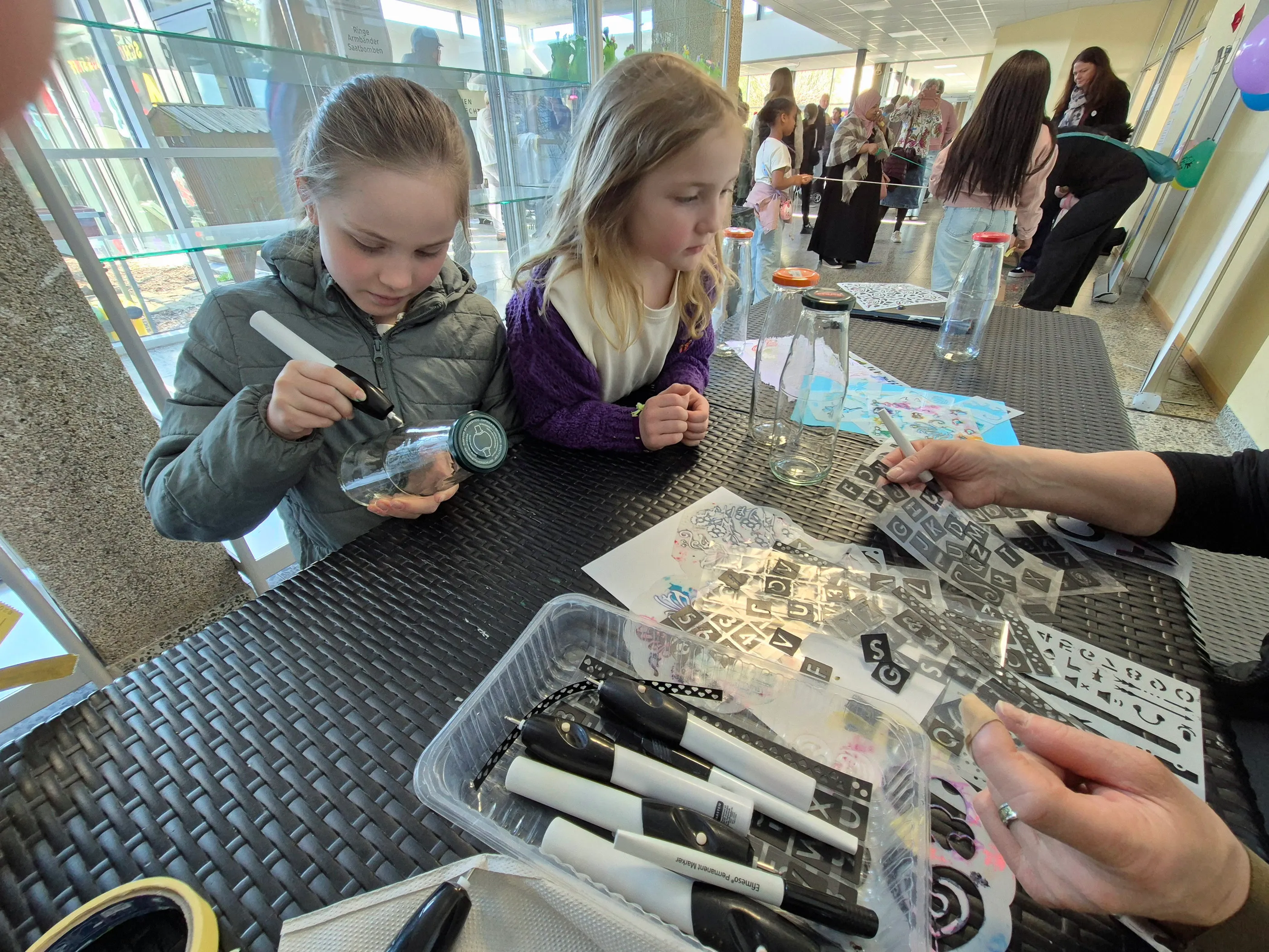 Zwei Mädchen bemalen mit Stiften und Schablonen Papier an einem Tisch, während eine erwachsene Person hilft. Im Hintergrund sind weitere Besucher eines Frühlingsfestes zu sehen.