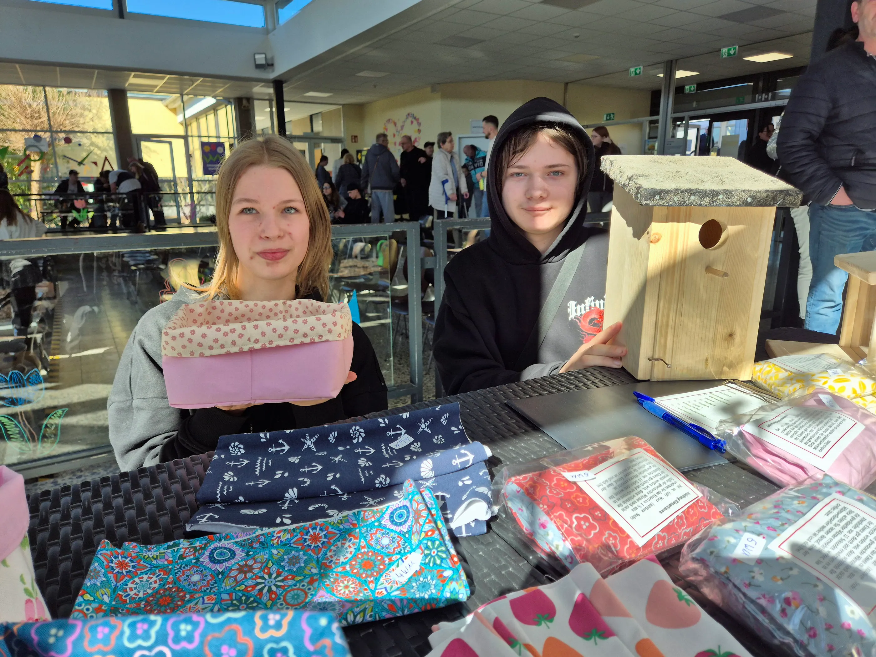 Zwei junge Menschen sitzen an einem Tisch mit selbstgemachten Artikeln, darunter Nähbeutel und Vogelhäuser, und präsentieren ihre Produkte auf einem Frühlingsfest der Phönixschule.