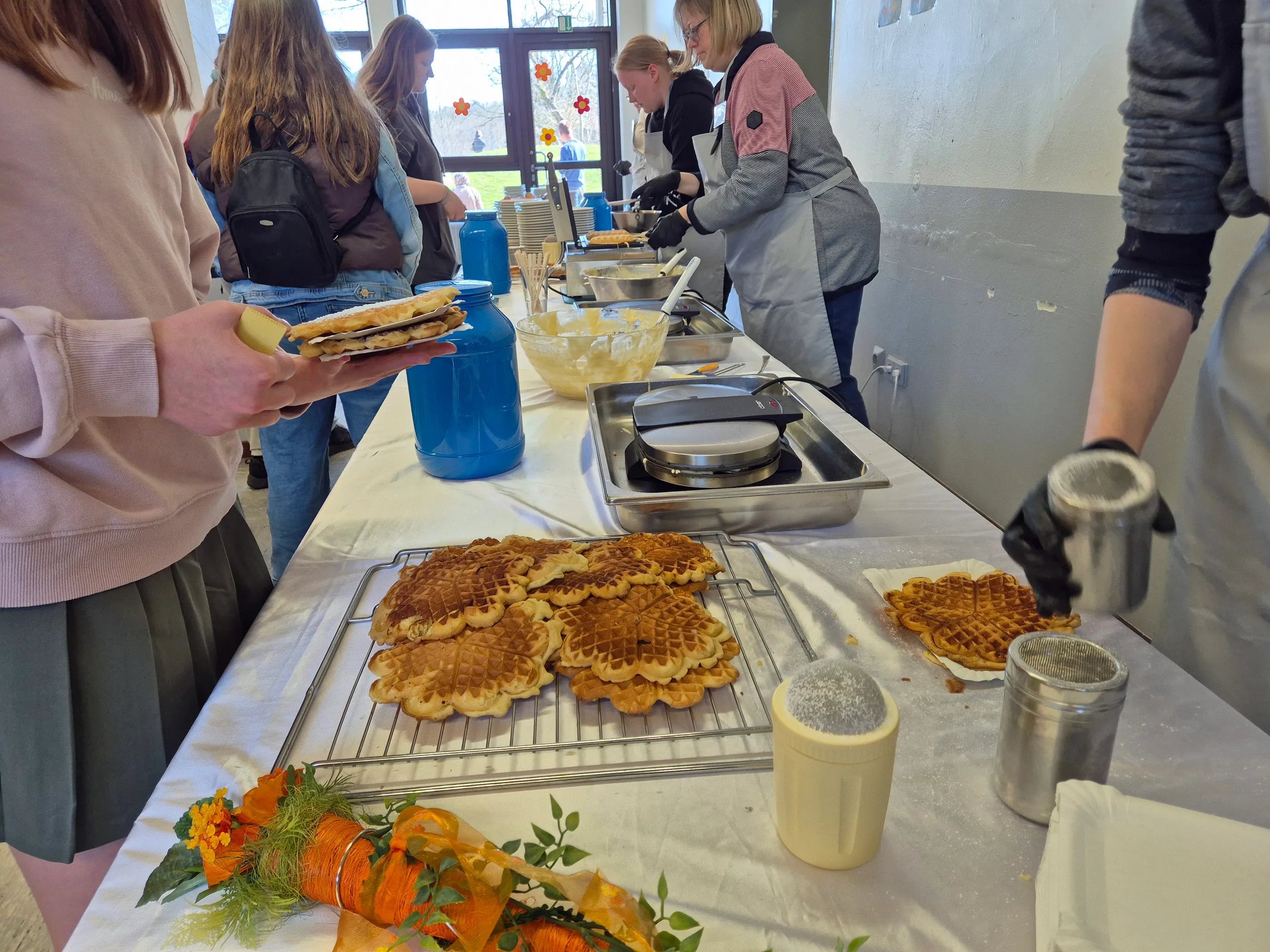 Ein Tisch mit vielen herzförmigen Waffeln und Streuseln steht im Mittelpunkt des Frühlingsfestes der Phönixschule. Im Hintergrund sind Menschen beim Zubereiten und Servieren zu sehen.