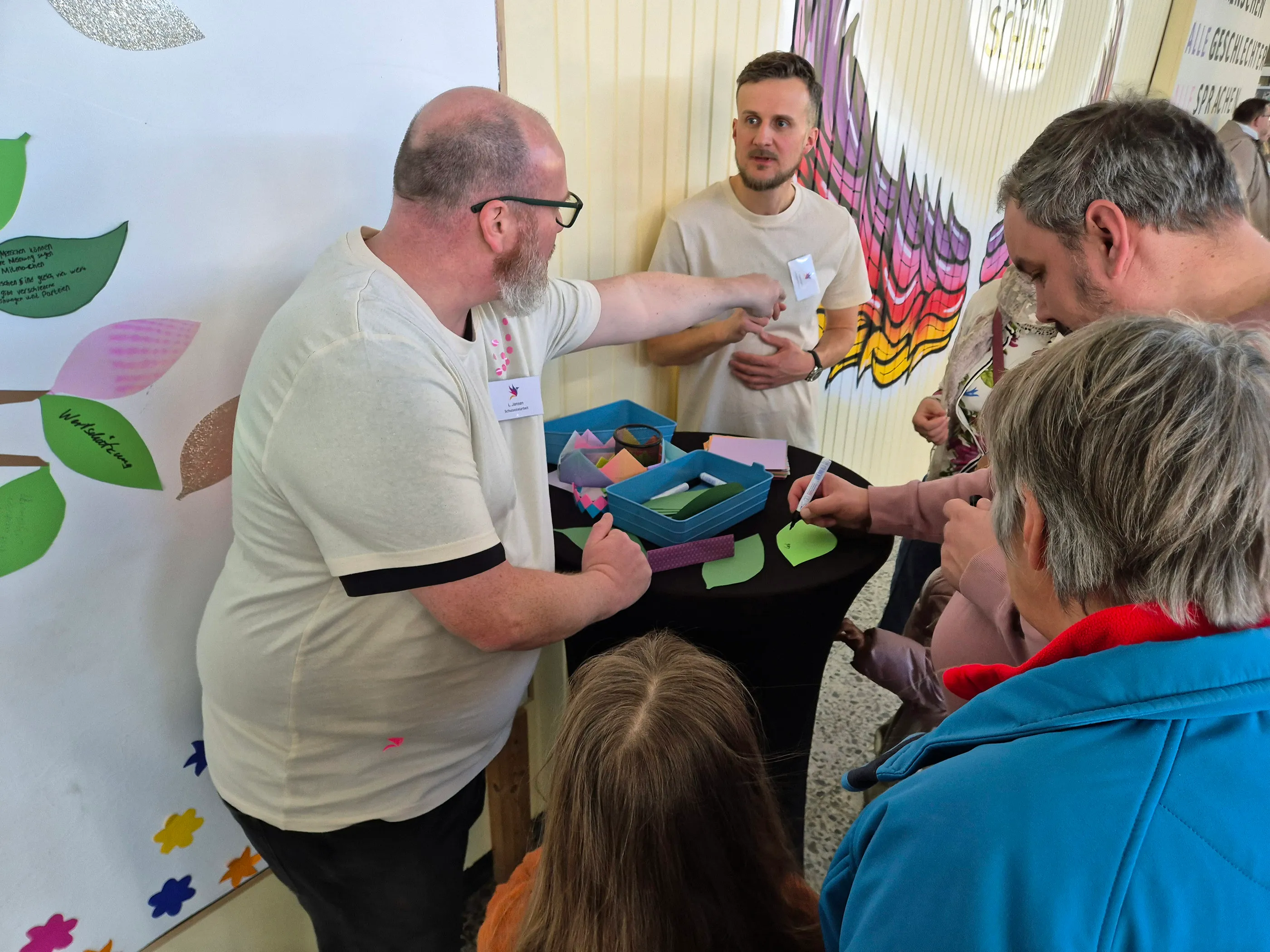 Zwei Männer und zwei ältere Frauen stehen um einen Tisch mit bunten Karten und schreiben Wünsche auf. Im Hintergrund eine Wand mit bunten Blättern und Schrift.