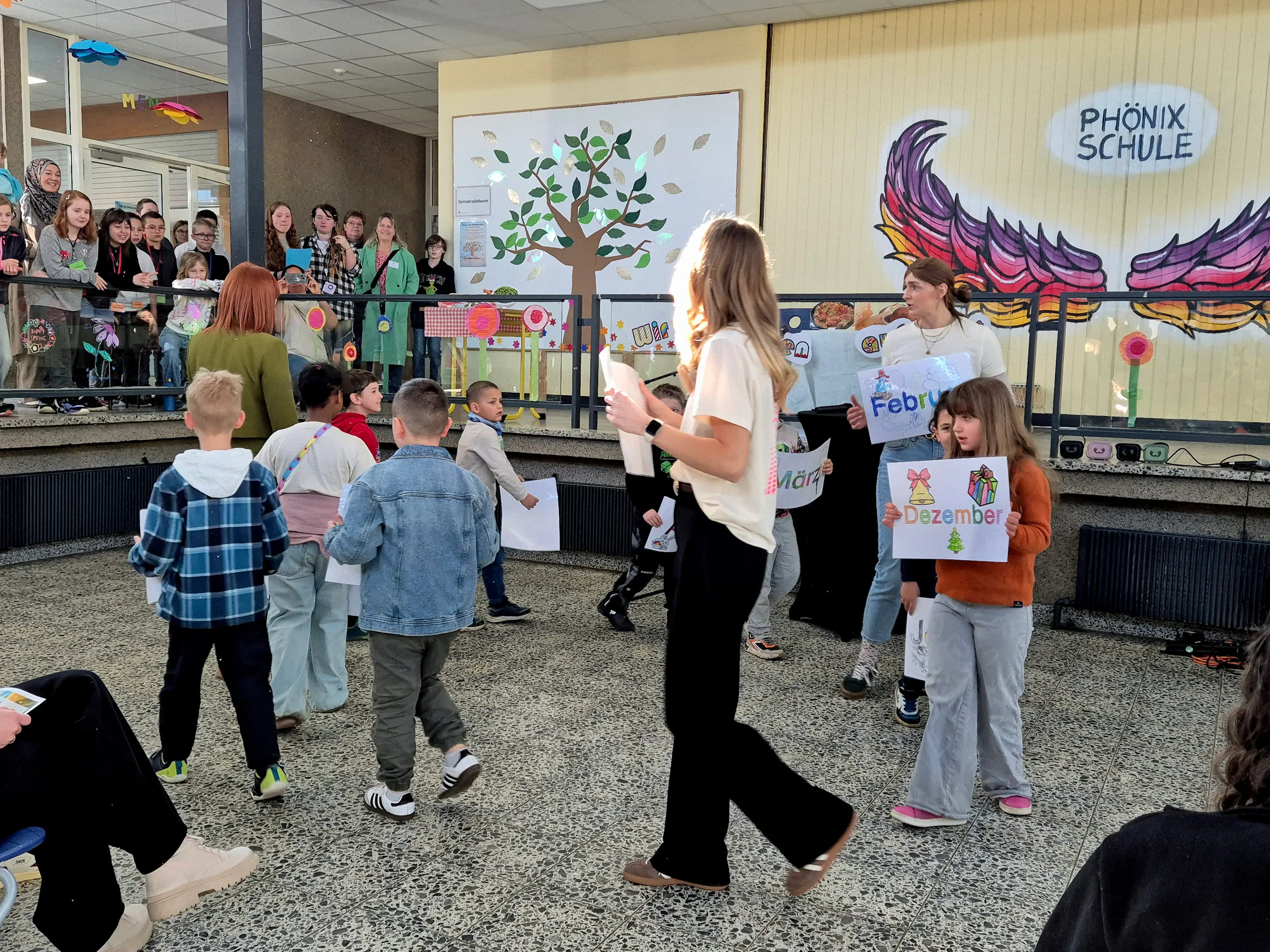 Eine Frau führt eine Gruppe von Kindern durch einen Raum, während andere Kinder und Erwachsene im Hintergrund zusehen. Die Kinder halten bunte Papiere mit Monatsnamen.