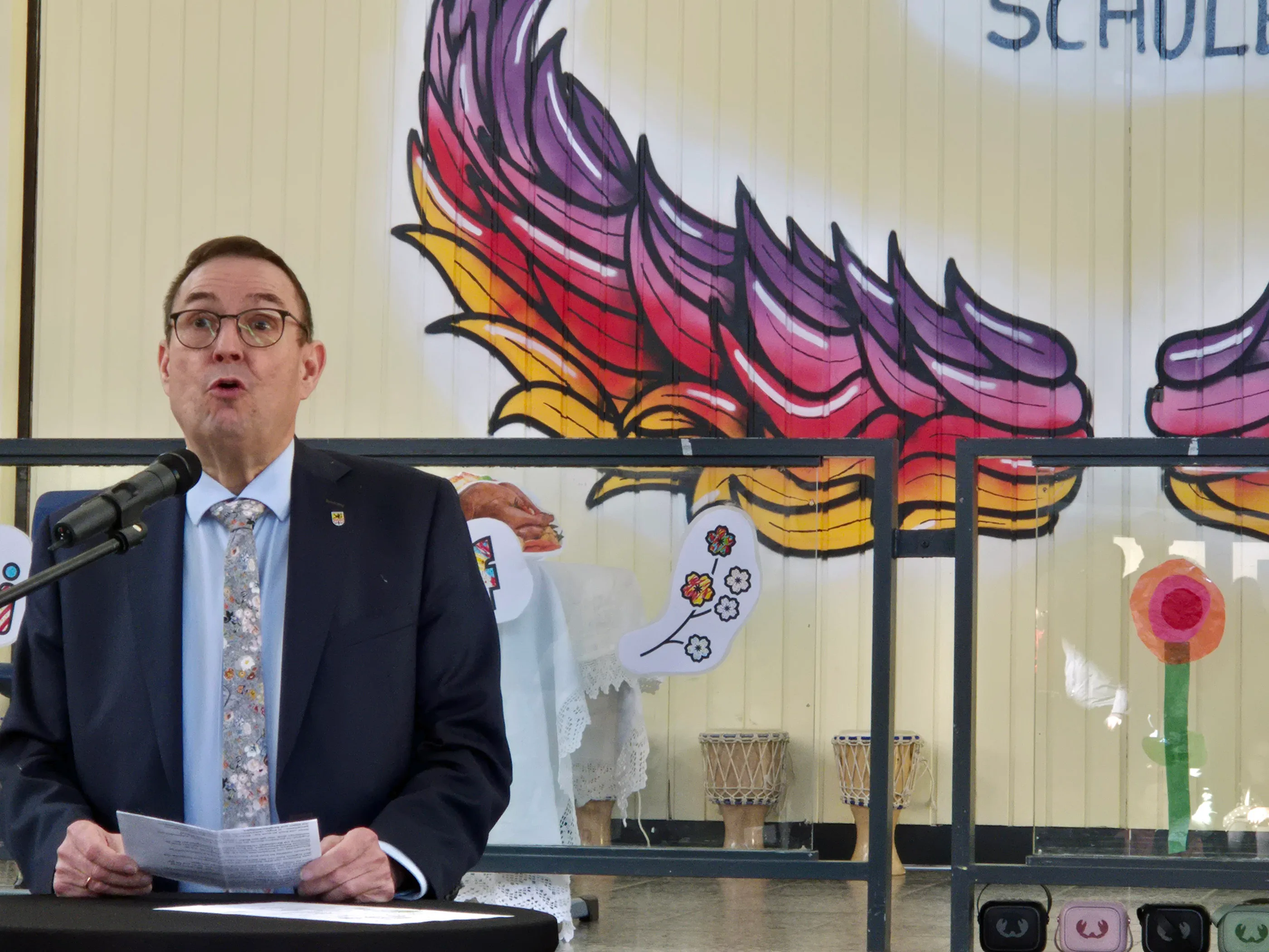 Ein Mann mit Brille hält eine Rede vor einem Mikrofon an einem Pult. Im Hintergrund ein farbenfrohes Wandbild mit einem stilisierten Phönix und Blumen.