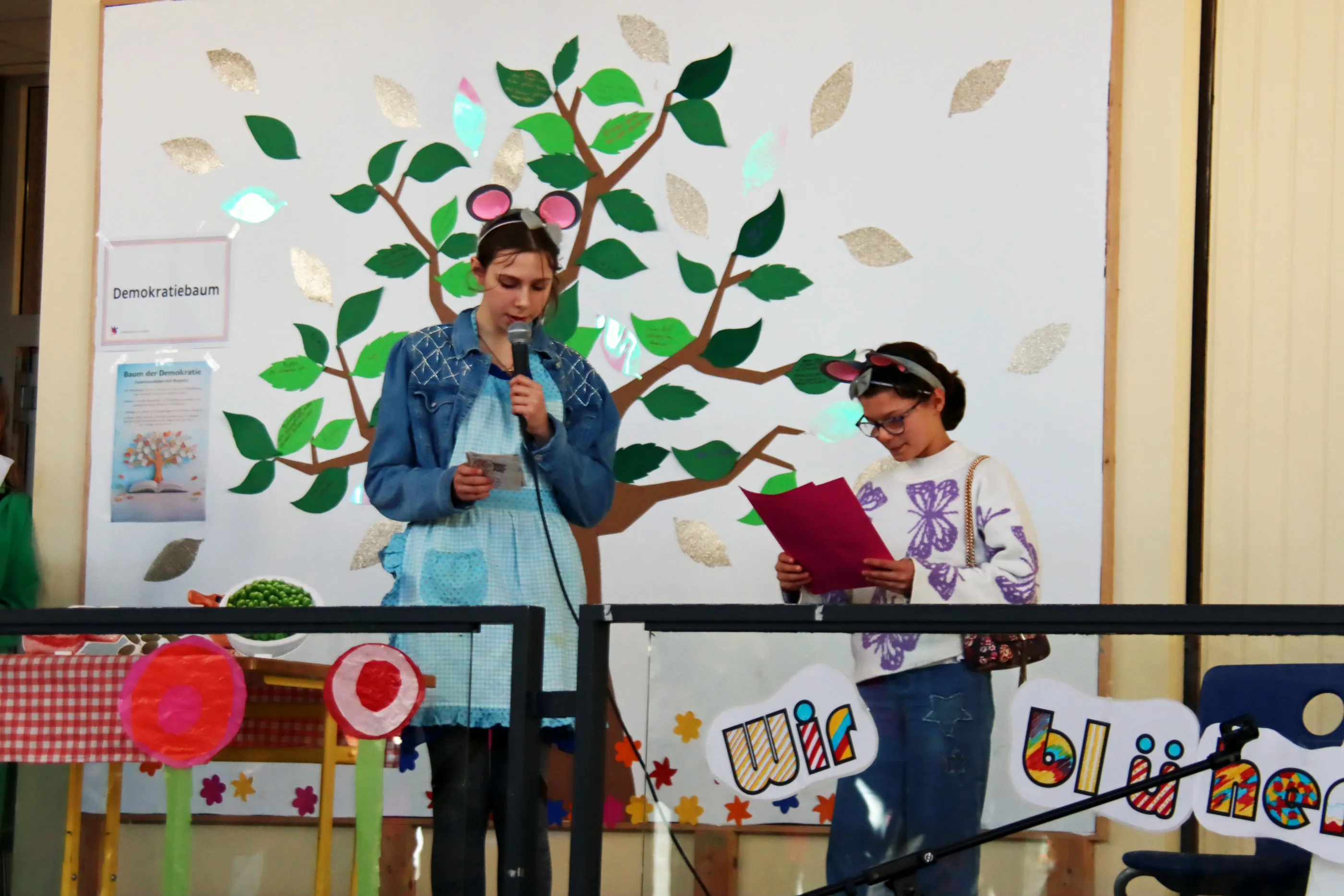 Zwei Mädchen halten Reden vor einem selbstgemalten Baum, der die Demokratie symbolisiert. Im Hintergrund bunte Dekorationen und das Motto des Frühlingsfestes: „Wir blühen auf“.
