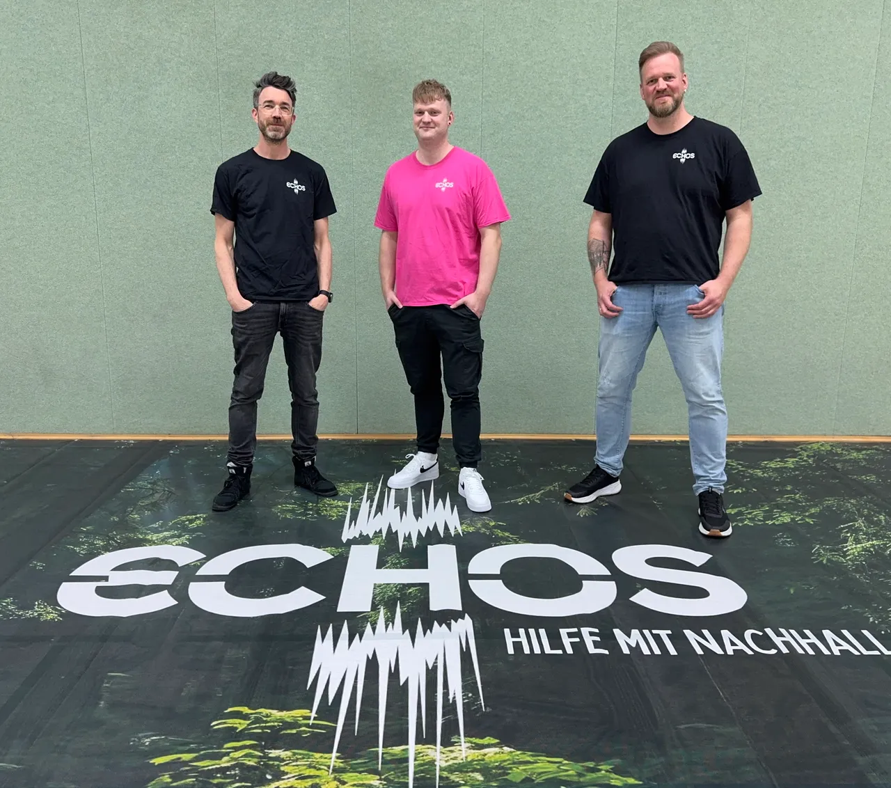 Drei Männer stehen vor einem grünen Hintergrund mit dem Schriftzug „ECHOS – HILFE MIT NACHHALL“. Von links nach rechts: Christian Schmitt, Hendryk Eckert und Gerrit Eckert, die Initiatoren der Benefizveranstaltung „Echos“.