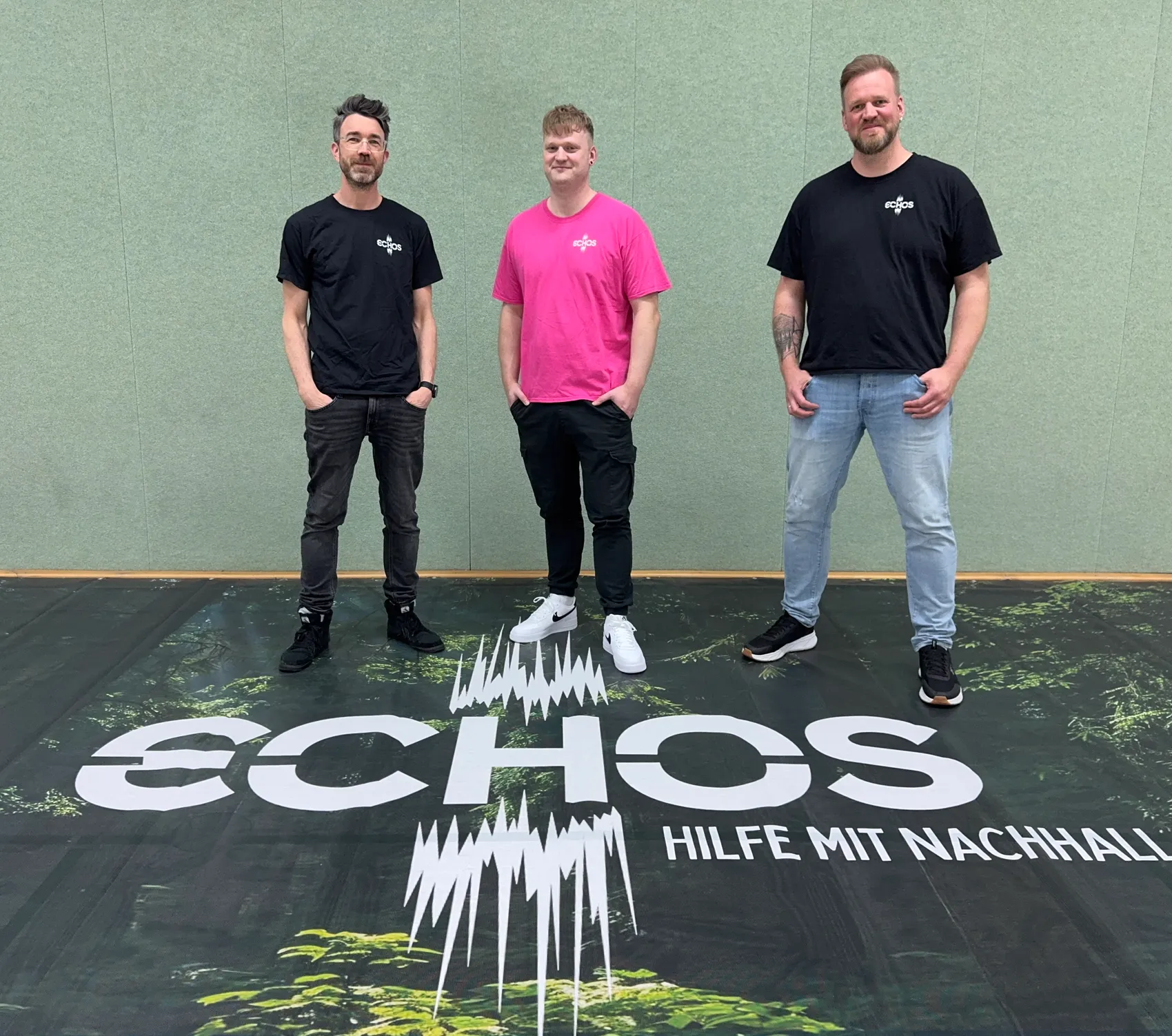 Drei Männer stehen vor einem grünen Hintergrund mit dem Schriftzug „ECHOS – HILFE MIT NACHHALL“. Von links nach rechts: Christian Schmitt, Hendryk Eckert und Gerrit Eckert, die Initiatoren der Benefizveranstaltung „Echos“.