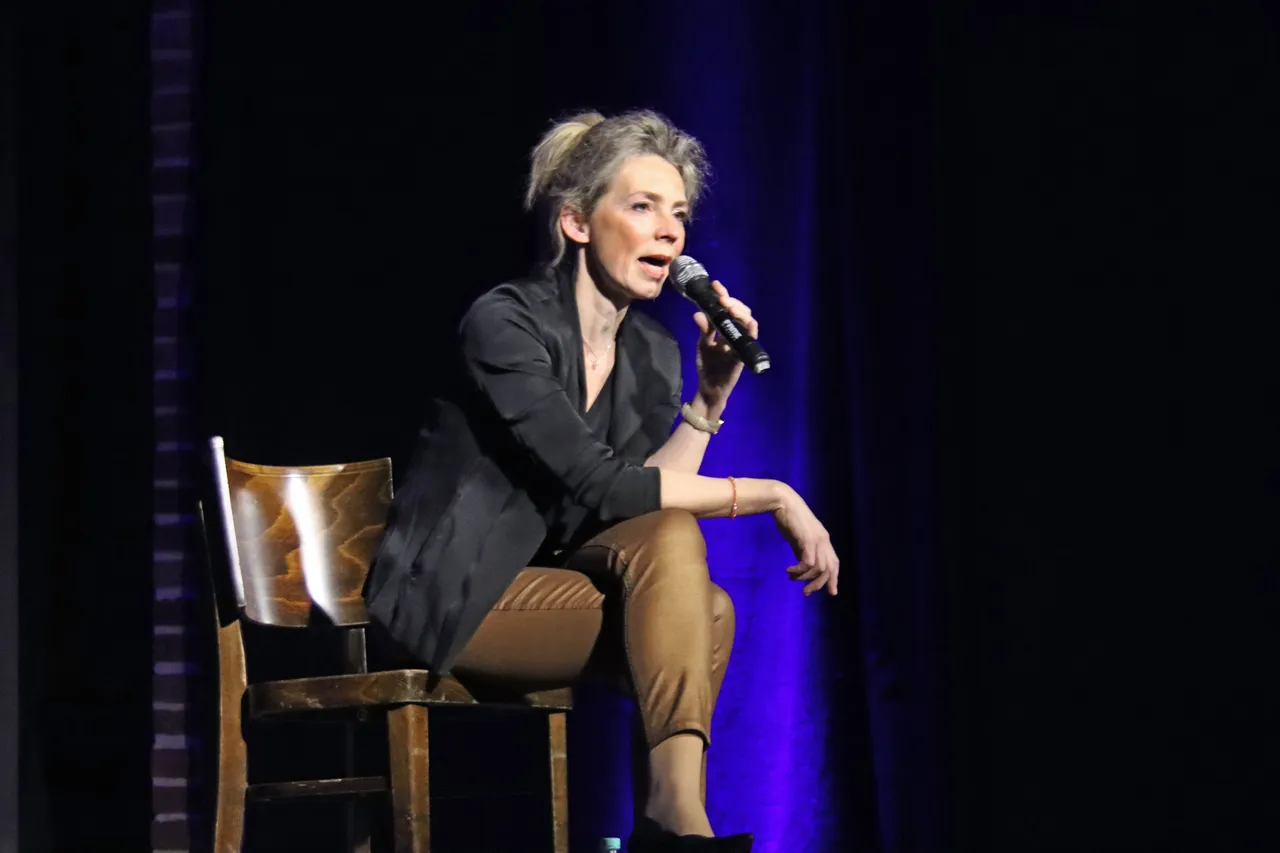 Christine Schütze sitzt auf einem Stuhl auf der Bühne und spricht in ein Mikrofon, während sie eine Performance gibt.