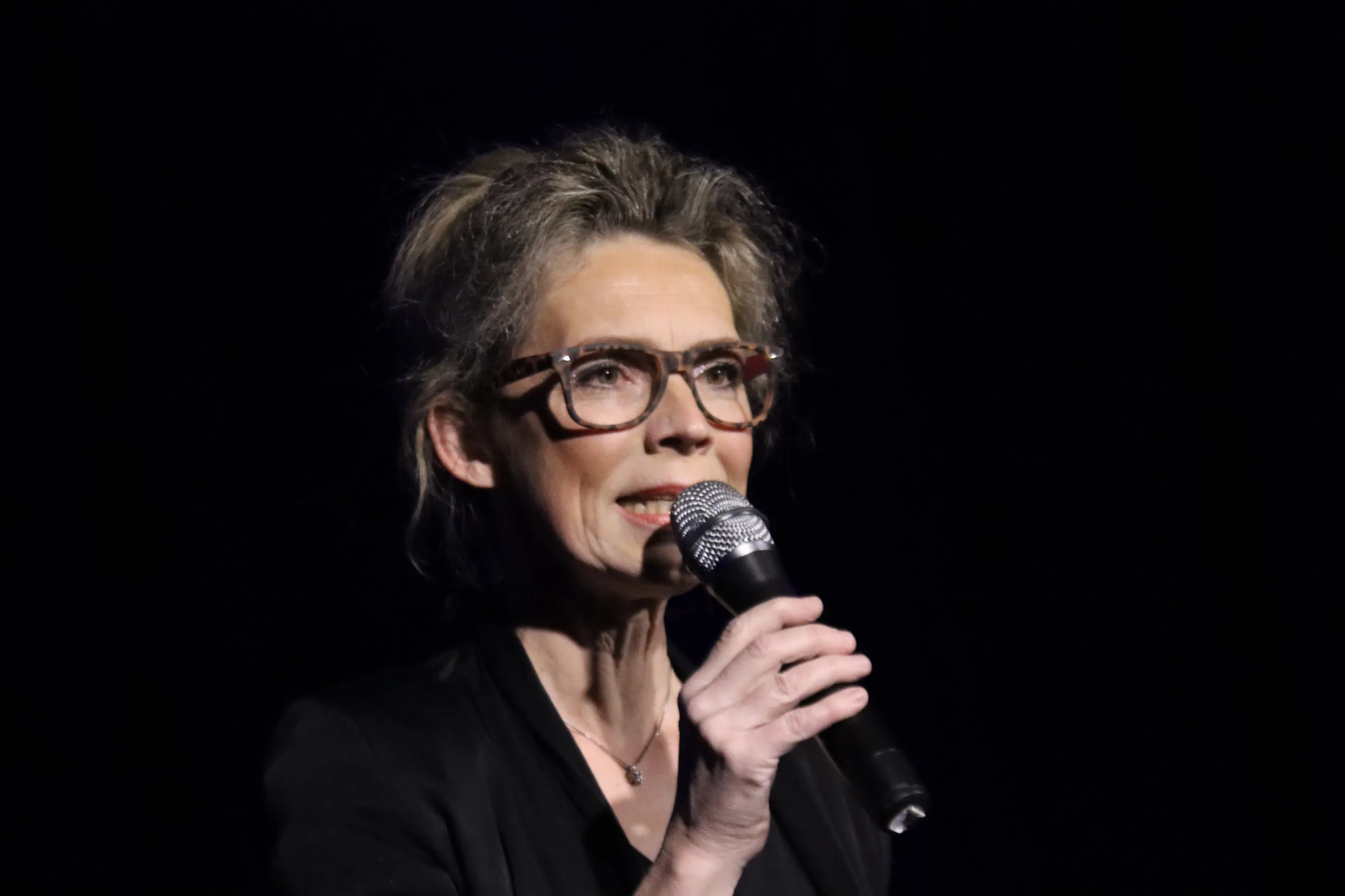 Christine Schütze hält ein Mikrofon und spricht auf einer Bühne vor dunklem Hintergrund.