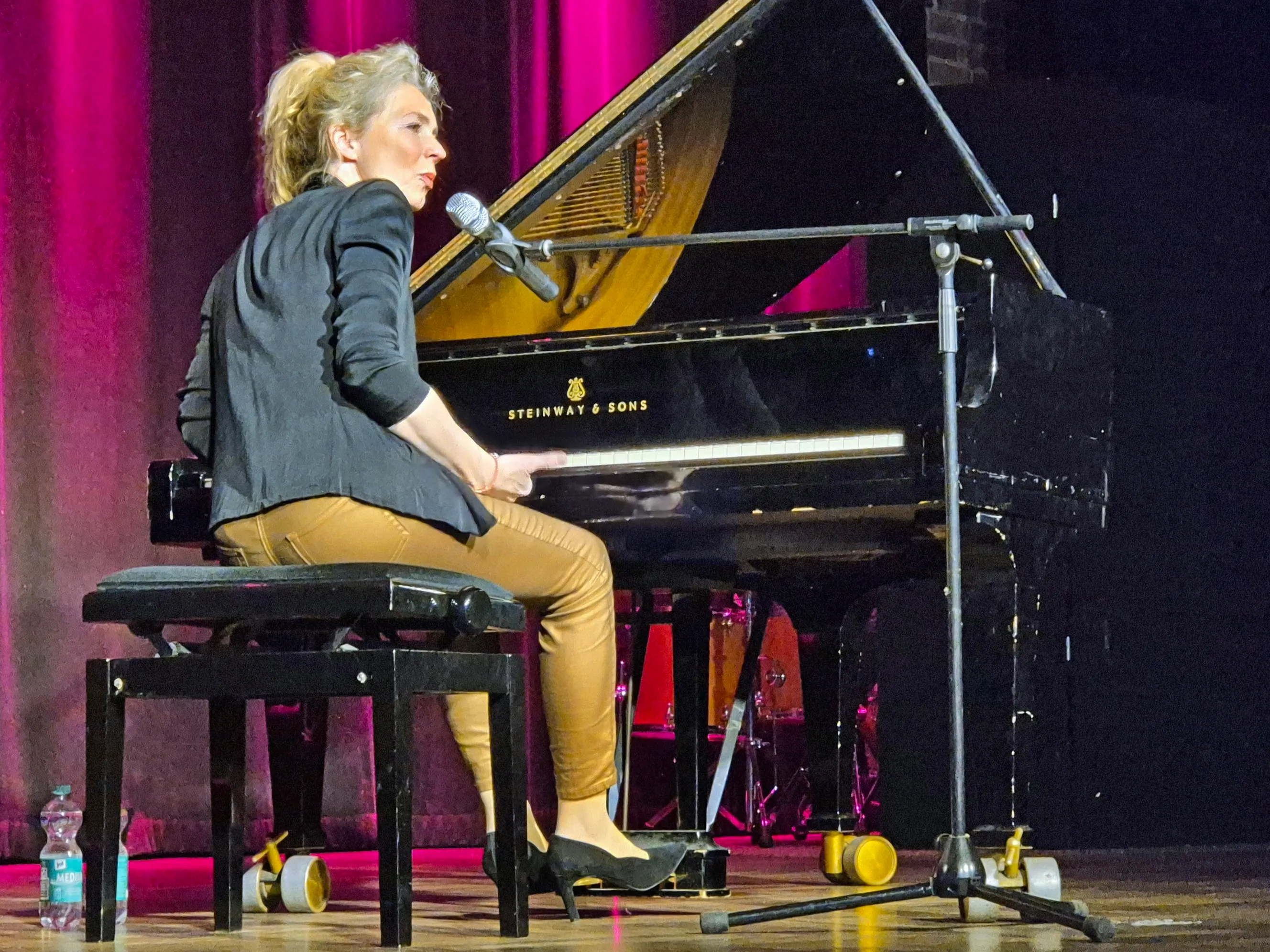 Christine Schütze sitzt am Flügel und singt in ein Mikrofon. Im Hintergrund ein roter Vorhang und Scheinwerfer.