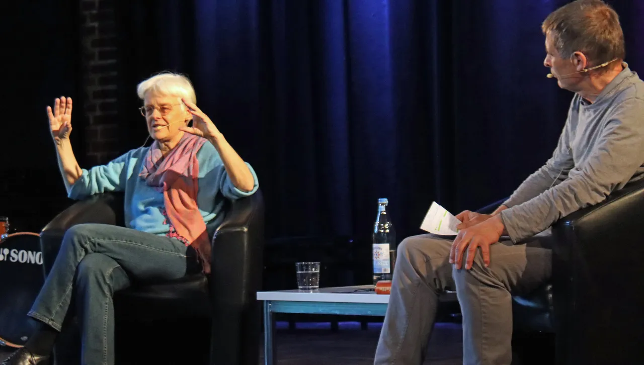 Ulrike Herrmann gestikulierend während einer Lesung, neben ihr ein Mann mit Notizen. Im Hintergrund ein dunkler Vorhang und ein Tisch mit Getränken.