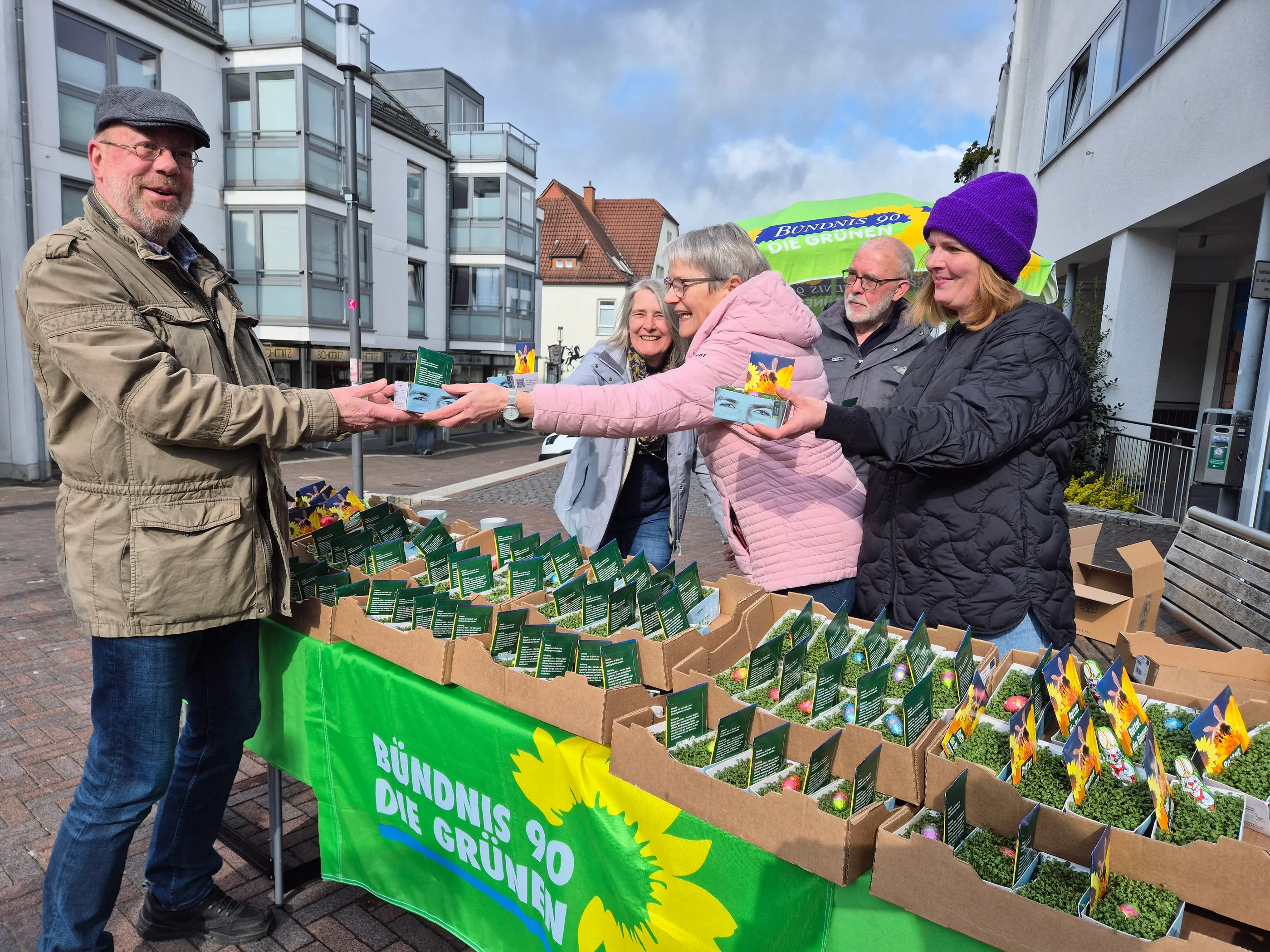 Ein Mann übergibt einer Frau an einem Stand mit Kresse und Blumensamen der Partei Bündnis 90/Die Grünen eine kleine Schale mit Kresse.