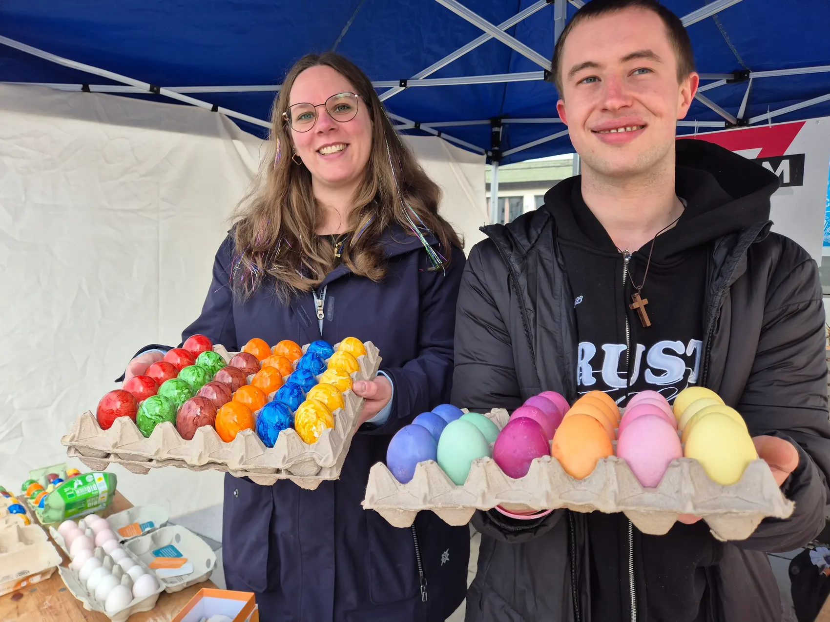 Zwei Personen halten Eierkartons mit bunt gefärbten Ostereiern vor sich. Sie stehen an einem Stand unter einem blauen Pavillon, im Hintergrund sind weitere Eier und ein Schild zu sehen.