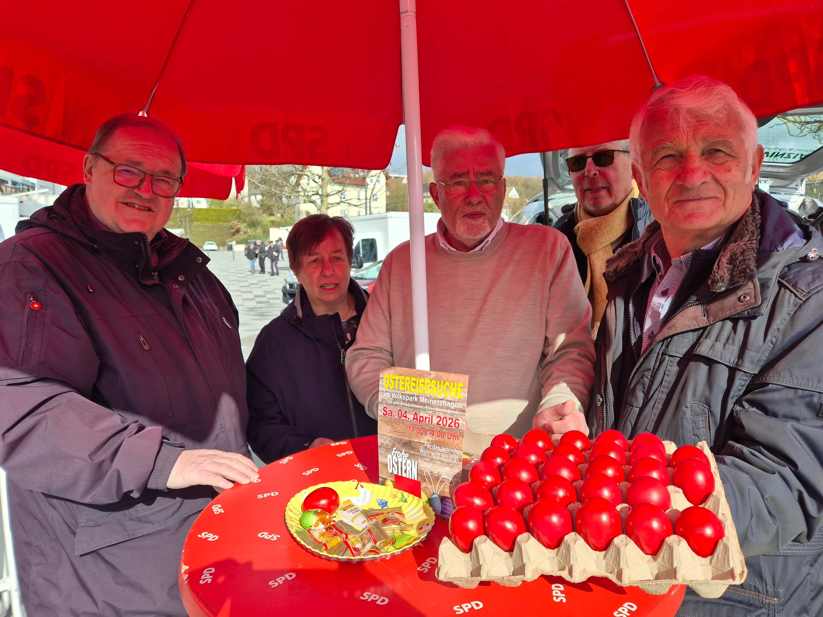 Drei SPD-Politiker posieren lächelnd mit einer Schachtel roter Ostereier und einem Kuchen mit SPD-Logo vor einem roten Sonnenschirm.